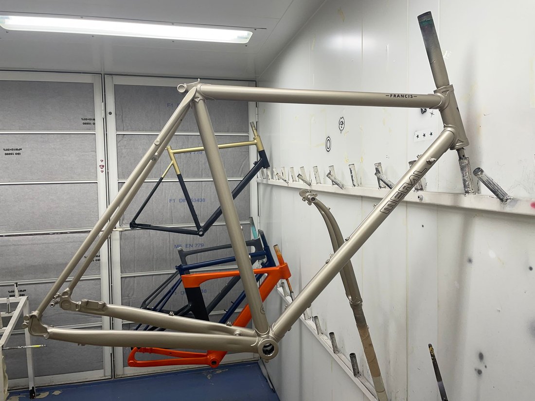 Cadre de vélo en acier, peint couleur champagne, suspendu dans une cabine de peinture.